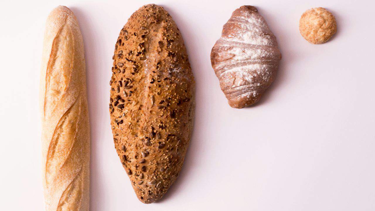 Four types of bread are arranged on a white surface: a baguette, a seeded oval loaf, a croissant, and a small round roll.
