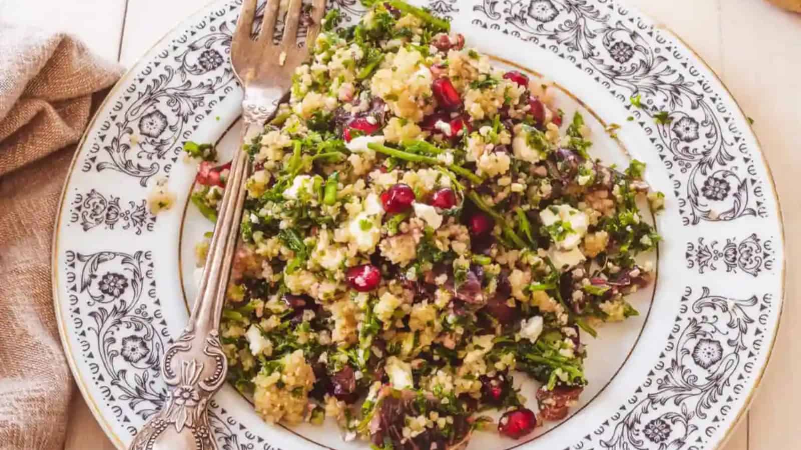 A plate of quinoa salad with broccoli, pomegranate seeds, nuts, and crumbled cheese, served with a fork on a decorative plate.