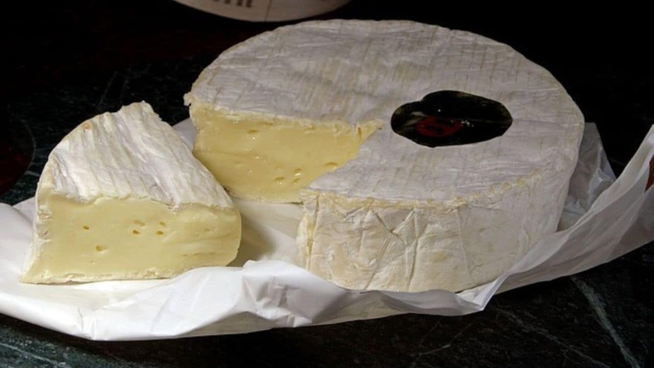 A wheel of soft cheese with a slice cut out, showing a creamy interior, on white paper.