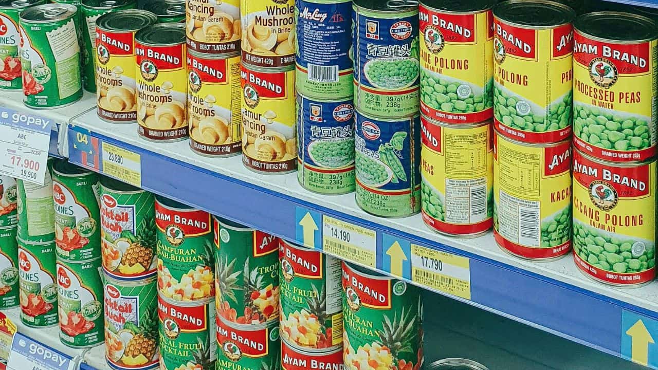 A supermarket shelf displays various canned foods, including brands of mushrooms, peas, beans, and mixed vegetables, with price tags visible below each item.