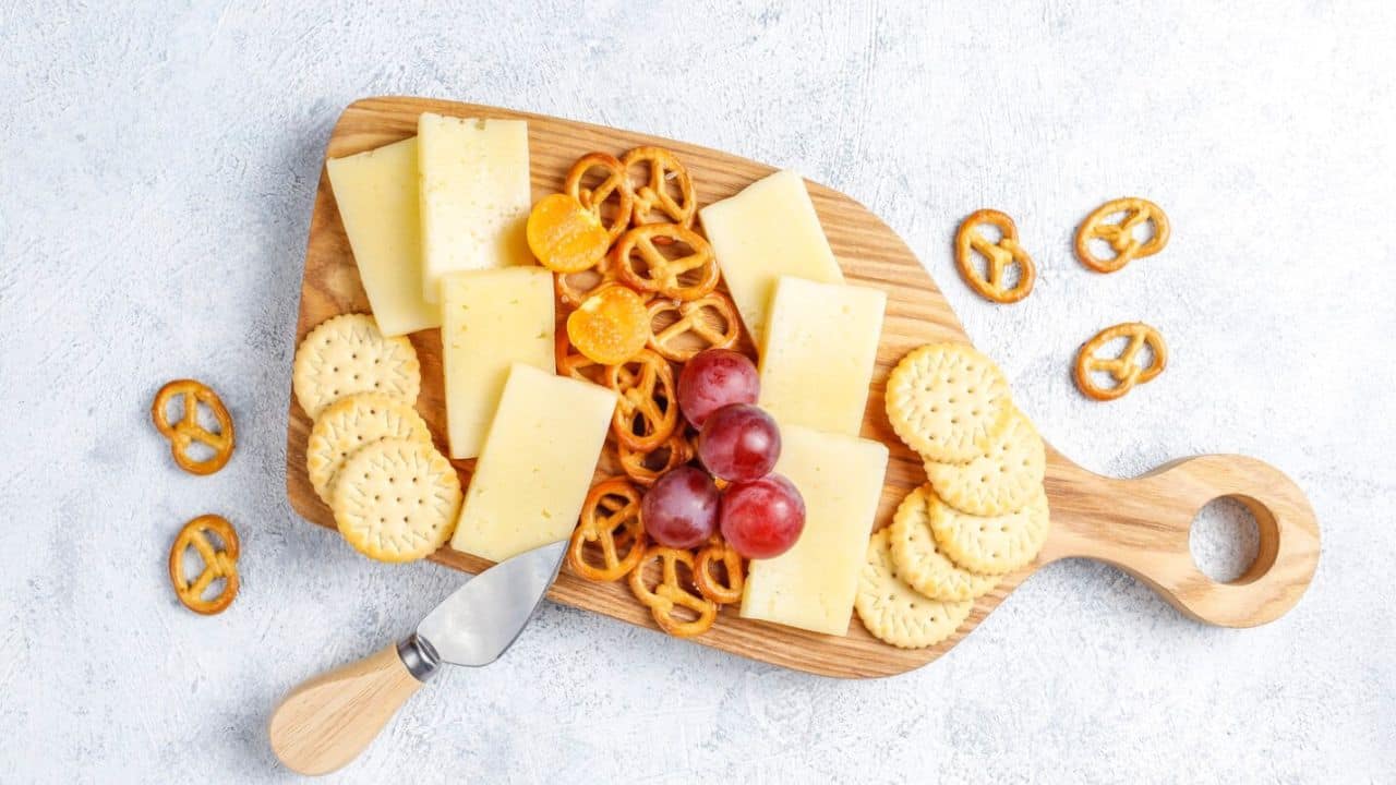 A wooden board with cheese slices, crackers, pretzels, grapes, and a cheese knife.
