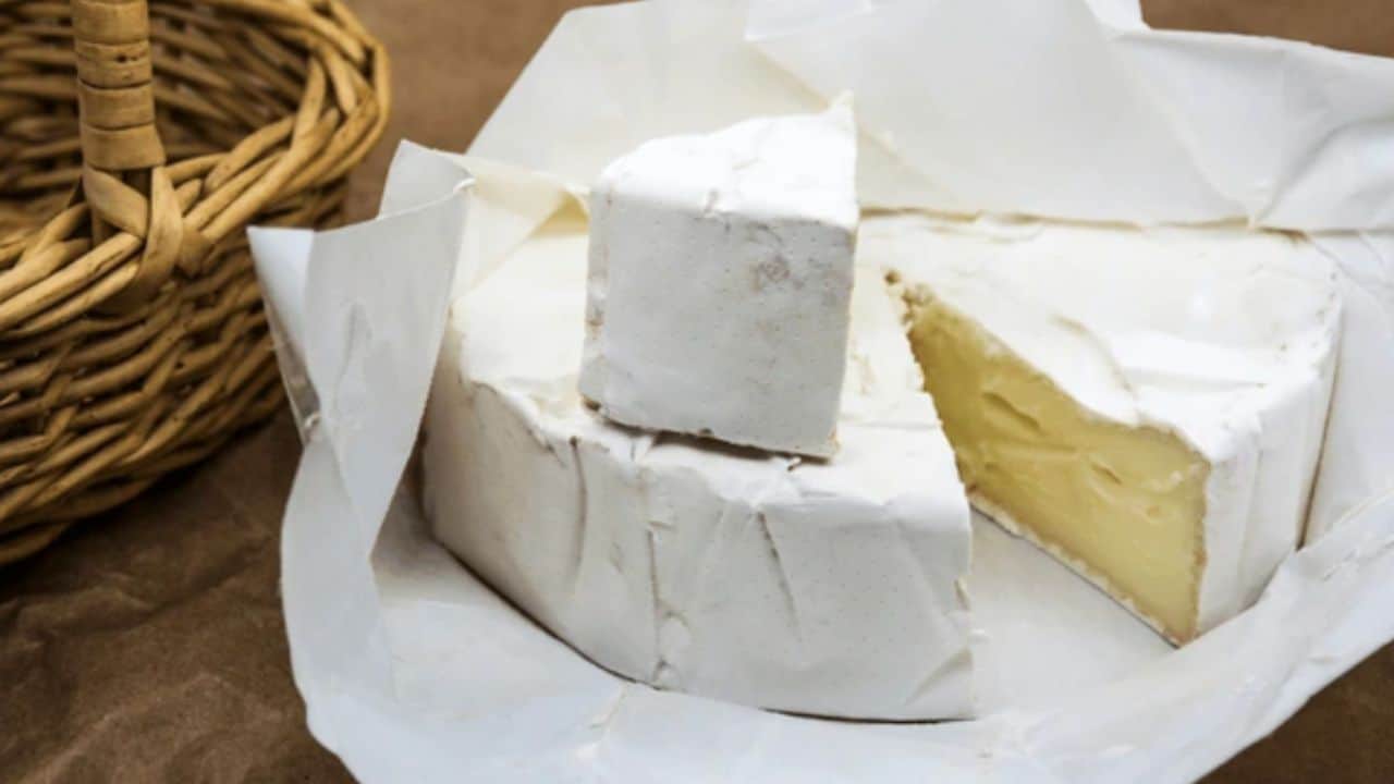 A wheel of soft cheese with a white rind is partially sliced open, with a wedge resting on top. A wicker basket is visible to the left.