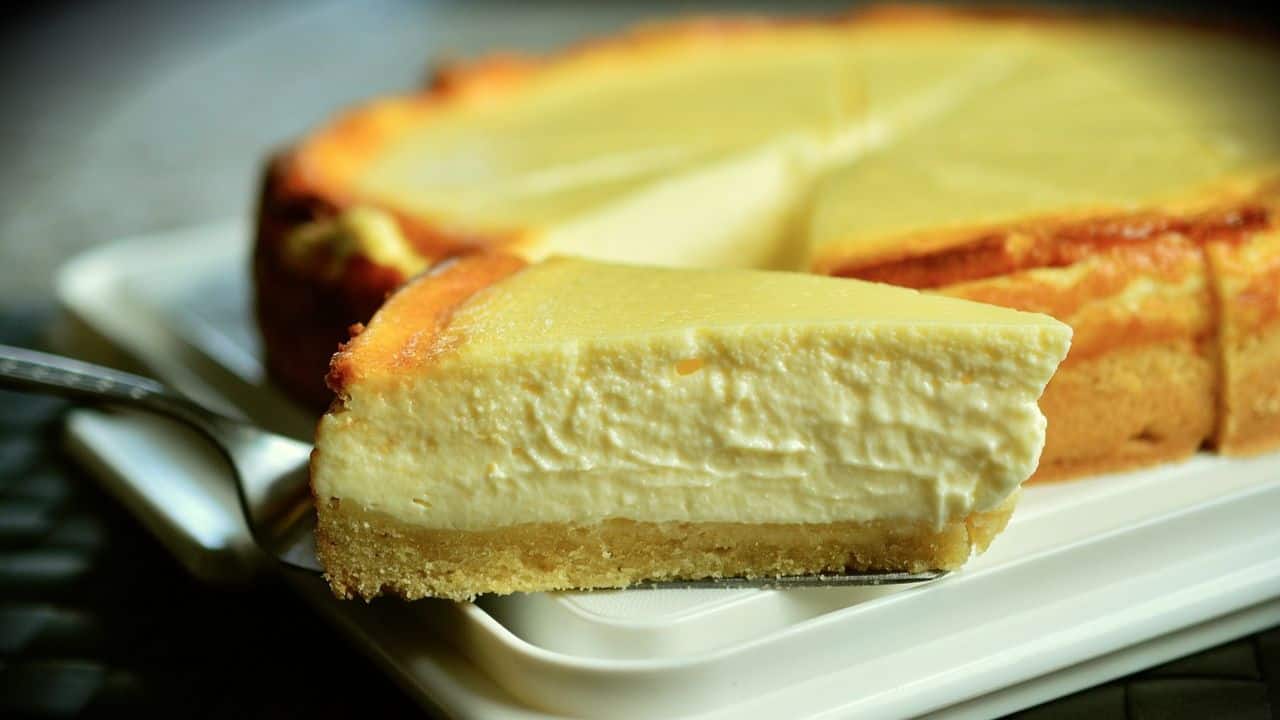 A slice of cheesecake with a creamy filling and a golden crust is lifted from a whole cheesecake on a white plate.
