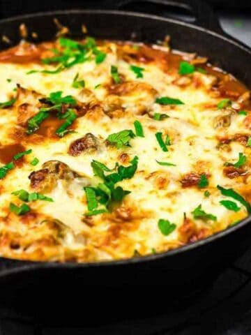 A skillet filled with cheesy baked dish topped with parsley, placed on a black wire rack, with a small bowl of shredded cabbage and parsley on the side.