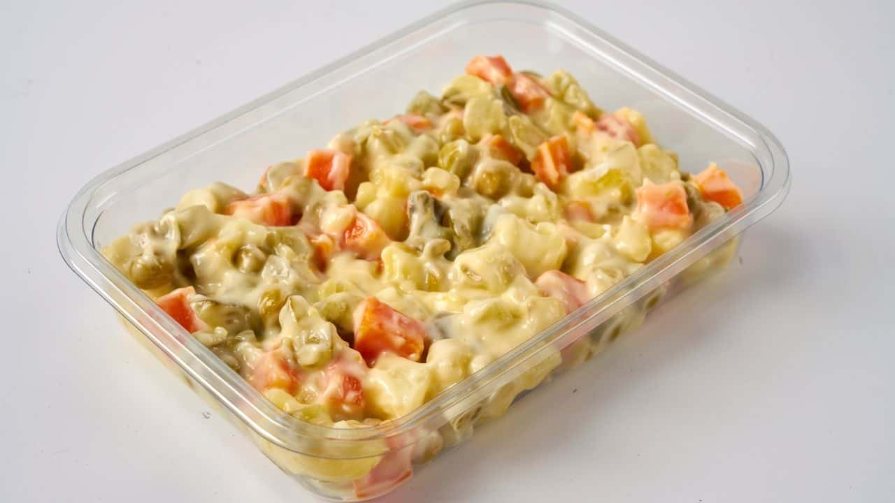 A clear plastic container filled with a creamy potato salad containing diced carrots, peas, and pickles on a white background.