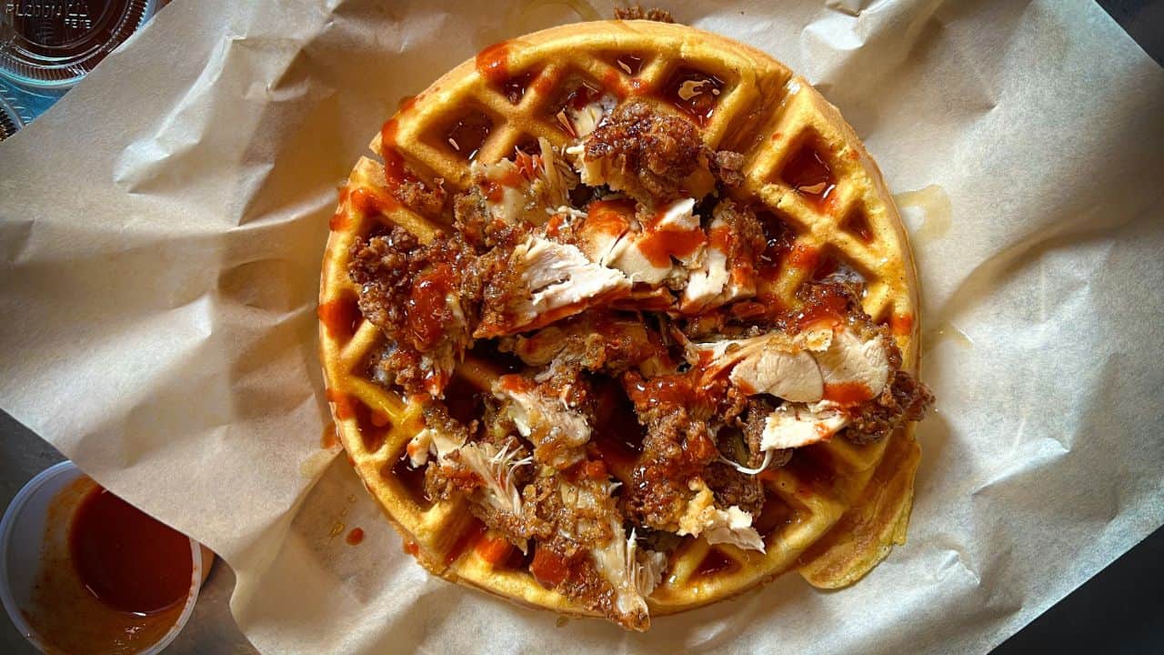A waffle topped with fried chicken pieces and drizzled with hot sauce, served on parchment paper.