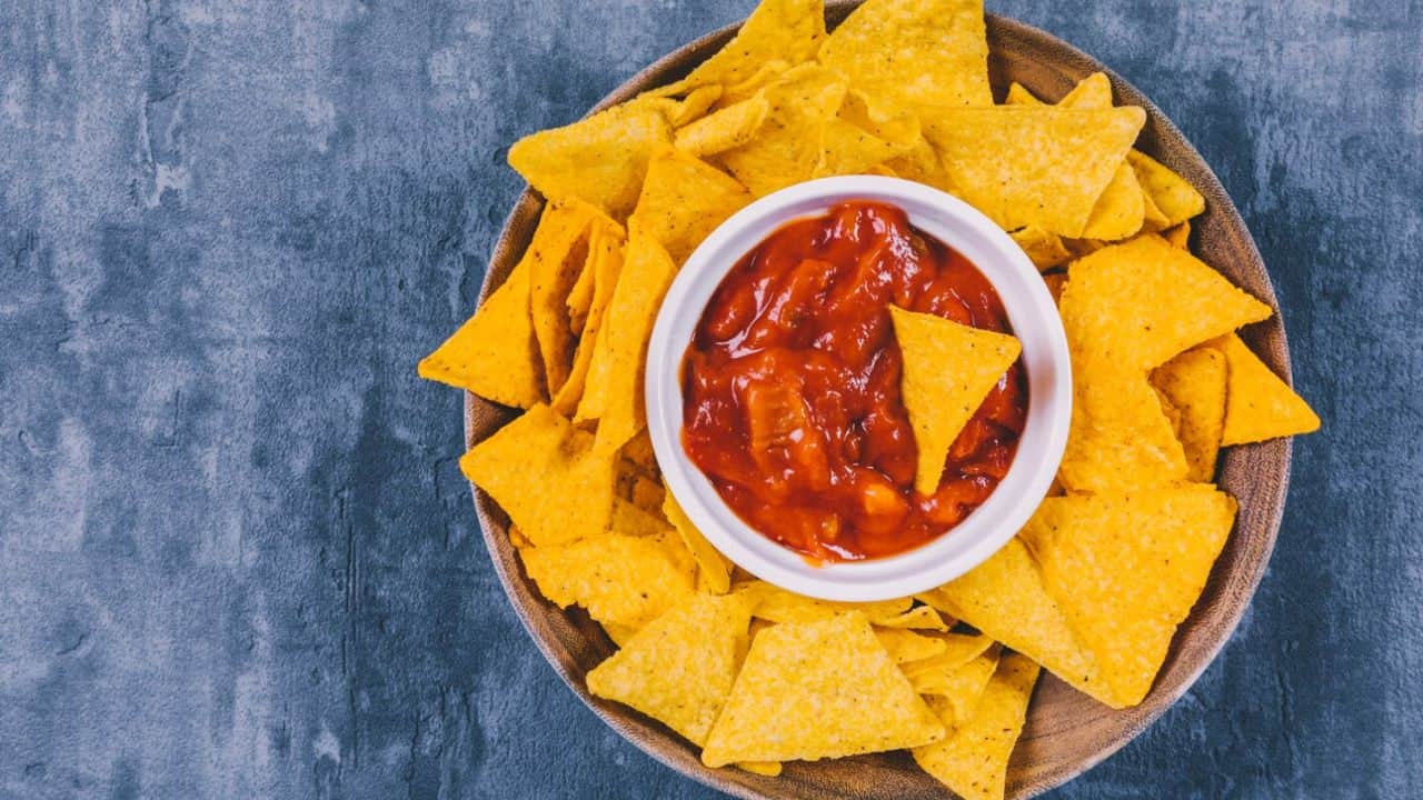 A bowl of triangular tortilla chips surrounds a smaller bowl of salsa on a dark textured surface.