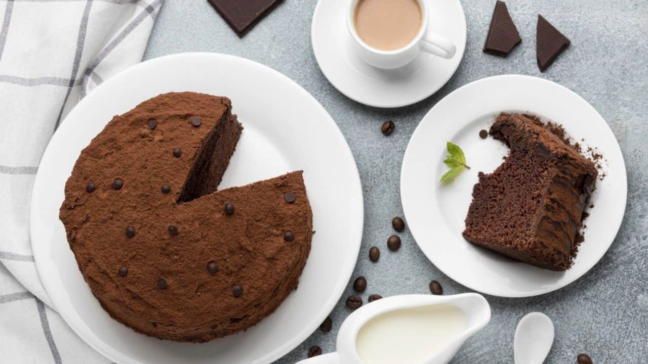 A chocolate cake with a slice missing sits on a plate next to a saucer with the slice, a cup of coffee, chocolate pieces, coffee beans, and a small pitcher of milk.