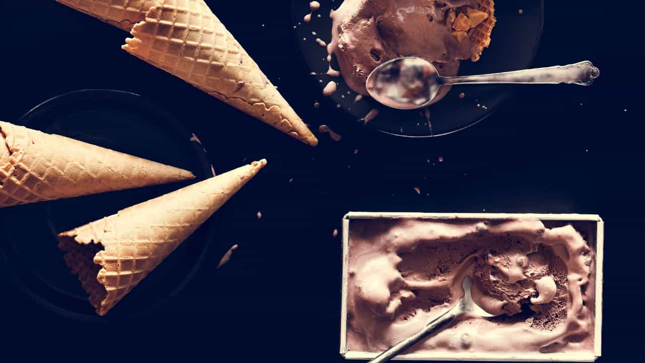 Ice cream cones and scoops of chocolate ice cream on a dark surface with spoons.