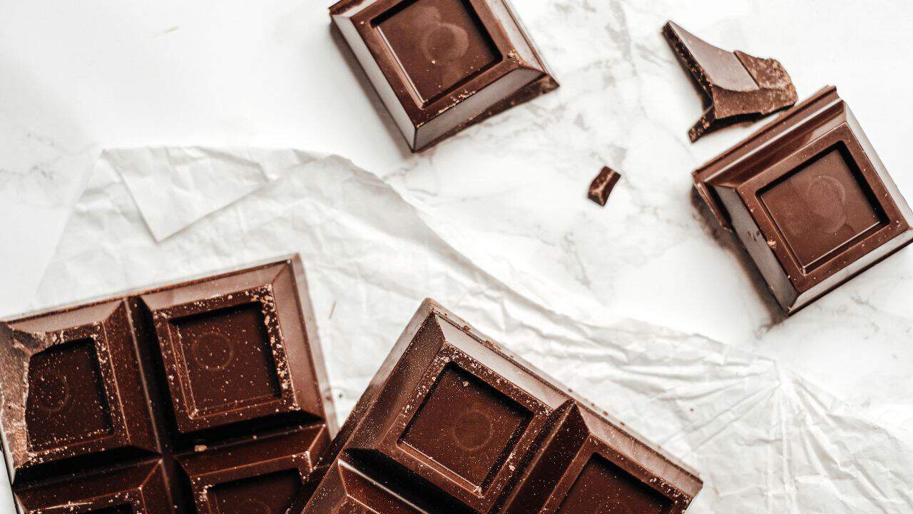 A bar of dark chocolate partially wrapped in white paper, with some pieces broken off and scattered around.