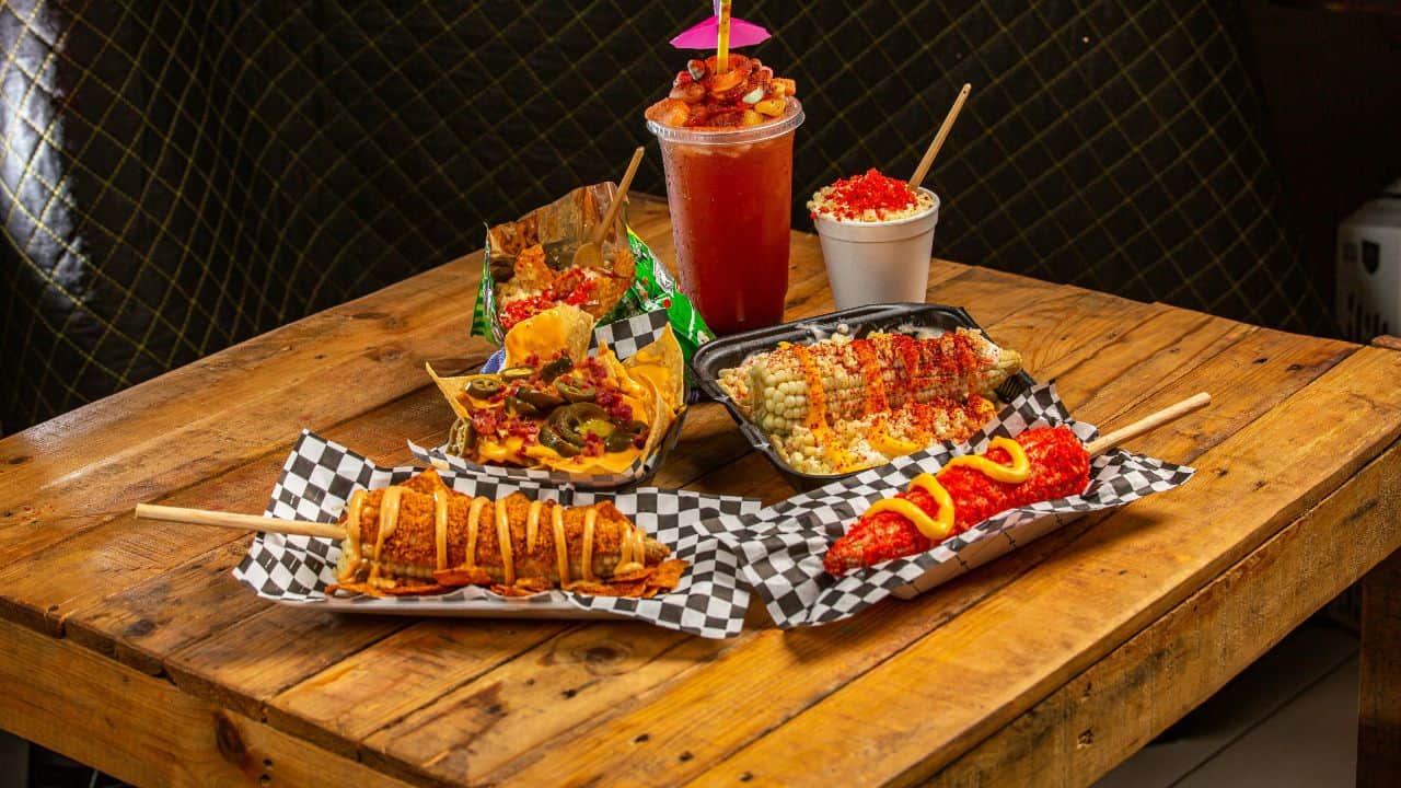 An assortment of Mexican street foods including elote, takis-covered corn, nachos, and a spicy drink on a wooden table.