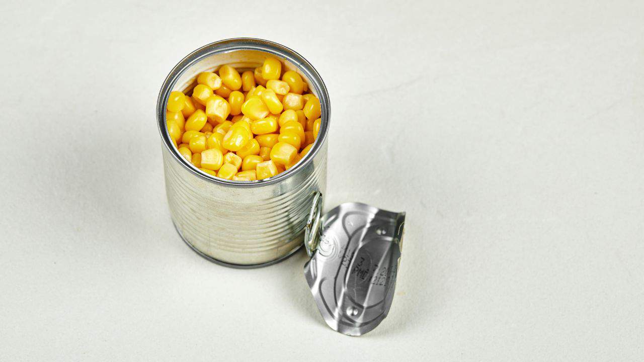 An opened can of corn with the lid partially detached, displaying yellow kernels on a light surface.