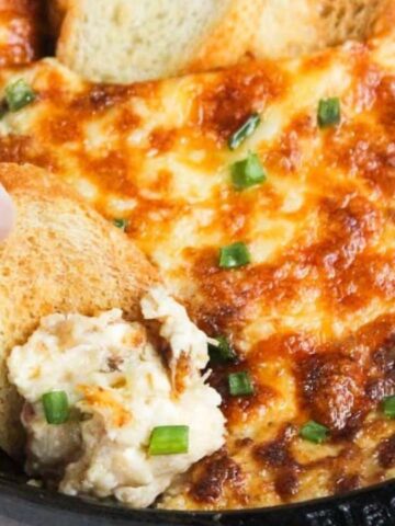 A hand dipping bread into a skillet of baked cheese and jalapeño dip.