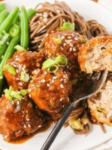 A plate with meatballs in sauce, garnished with sesame seeds and green onions, served with green beans and soba noodles. A fork holds a partly cut meatball.