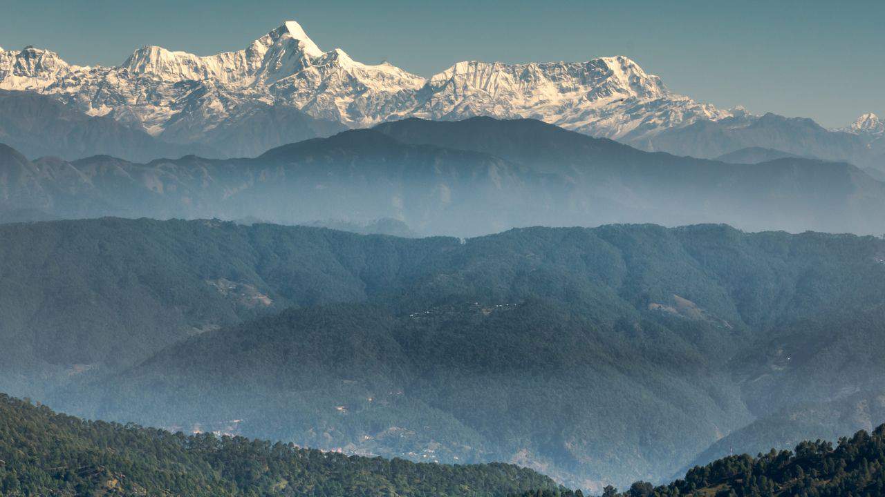 Snow-capped mountains with clear blue sky in the background, lush green forested hills and hazy atmosphere in the foreground.