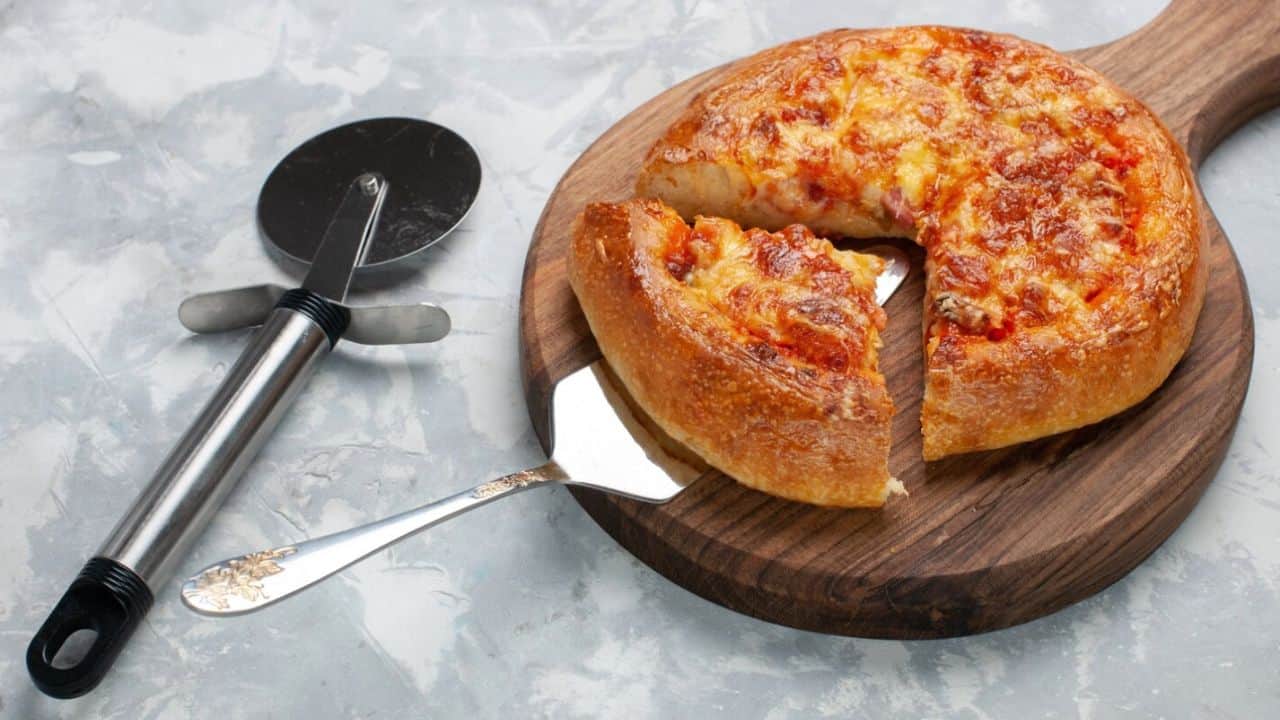 A deep-dish pizza with a slice cut out on a wooden board, alongside a pizza cutter.