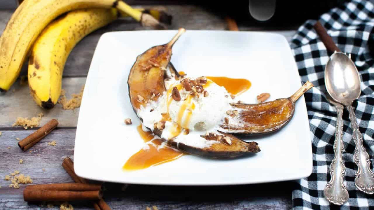 Grilled banana slices with vanilla ice cream, caramel sauce, and nuts on a white plate. Bananas, cinnamon sticks, and a checkered napkin are nearby.