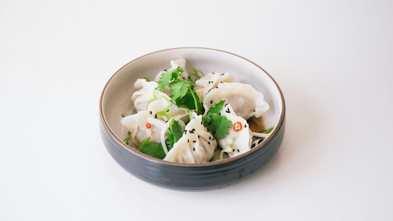 A bowl of dumplings garnished with cilantro, sesame seeds, and sliced red chili peppers on a white background.