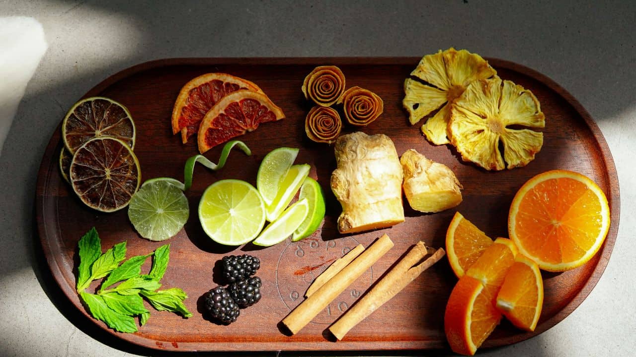 A wooden tray with assorted citrus slices, blackberries, ginger, cinnamon sticks, mint leaves, and fruit rolls arranged neatly under sunlight.