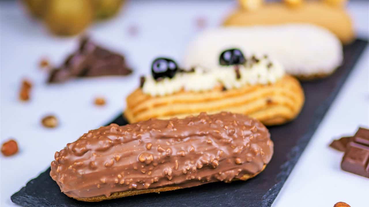 A slate tray holds a chocolate-coated éclair in the foreground, with two additional éclairs topped with cream and berries in the background. Scattered nuts and chocolate pieces surround them.