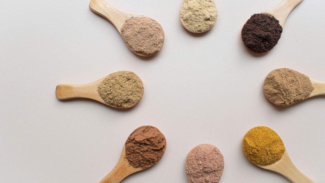 Seven wooden spoons displaying different colored powders arranged in a circular pattern on a light background.