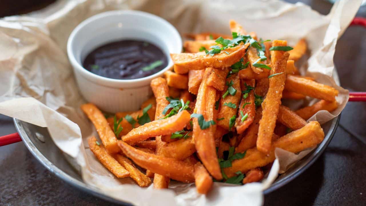 A bowl of sweet potato fries topped with herbs, served with a side of dipping sauce in a small white cup, placed on crumpled parchment paper in a round dish.