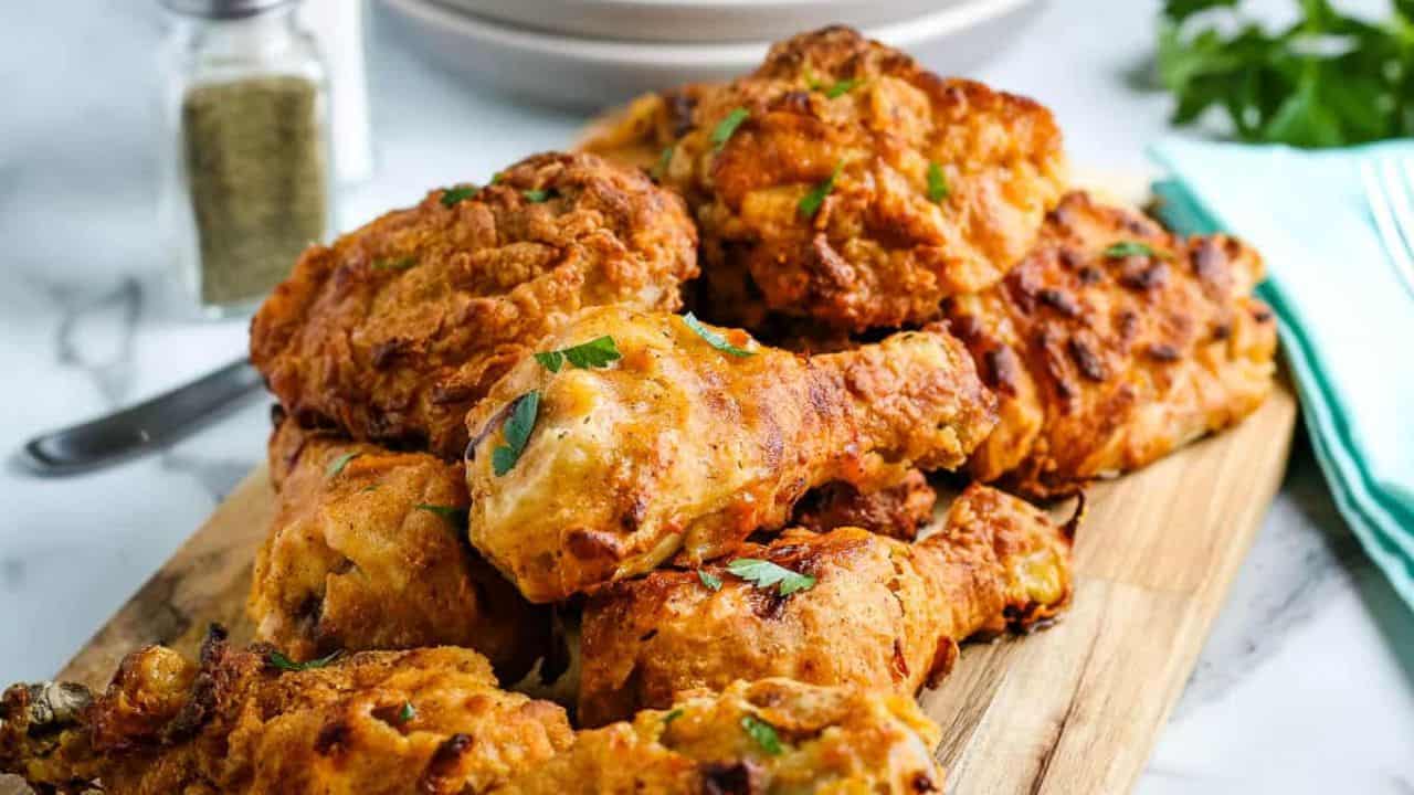 A pile of crispy fried chicken pieces garnished with parsley on a wooden board, with a jar of seasoning and stacked plates in the background.