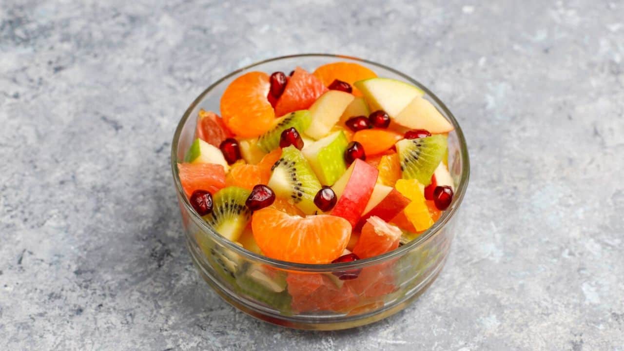 A glass bowl filled with a colorful fruit salad, including oranges, kiwi, apple pieces, grapefruit, and pomegranate seeds, placed on a gray textured surface.