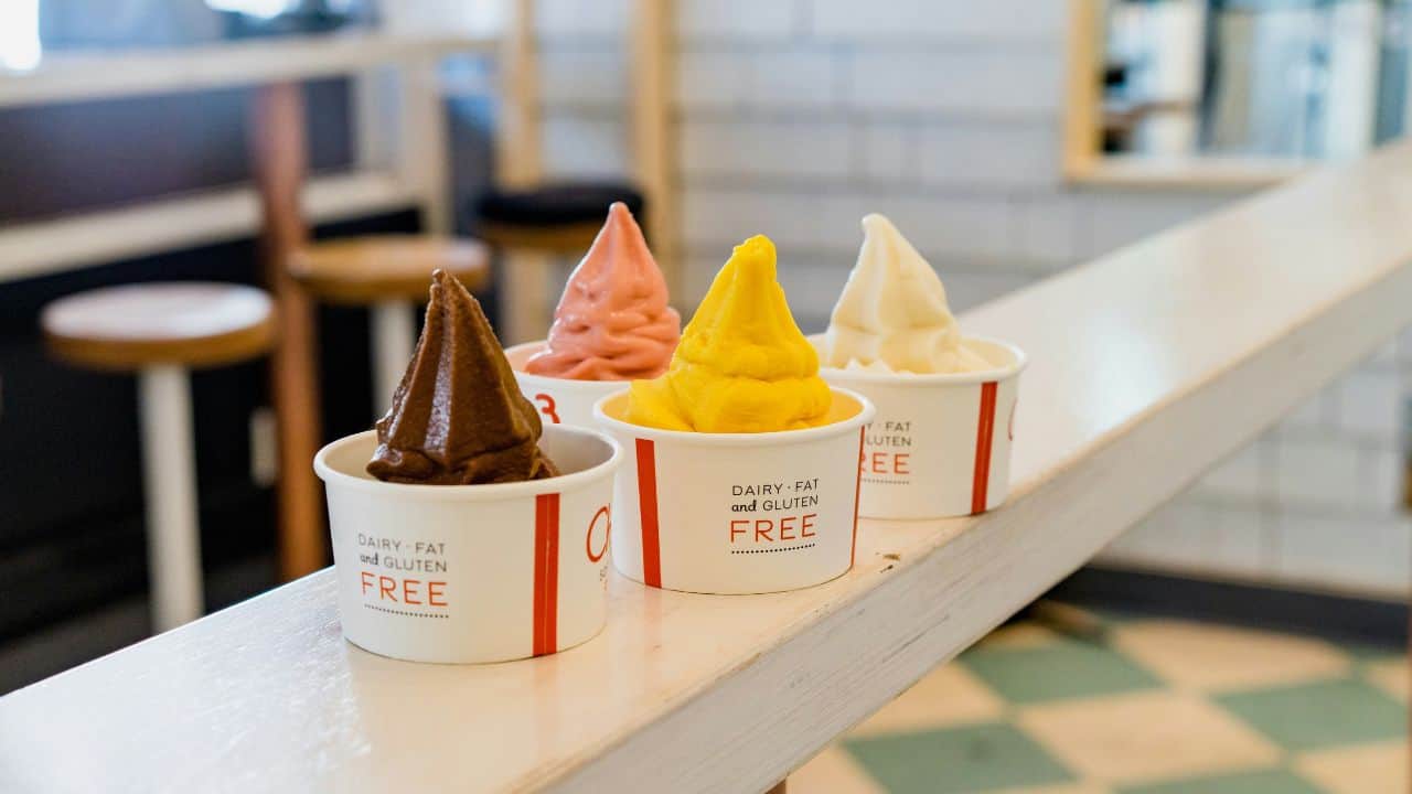 Four cups of dairy-free frozen dessert in chocolate, strawberry, mango, and vanilla flavors lined up on a wooden railing in a tiled room.