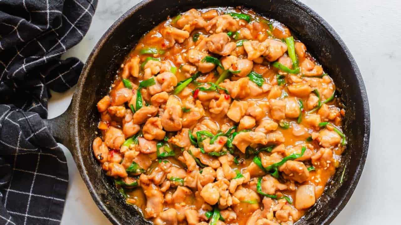 A skillet with cooked chicken pieces in a savory sauce, garnished with green onions. A checkered cloth is visible on the side.