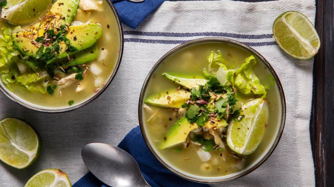 Two bowls of soup with avocado, lime, and herbs on a striped cloth. A spoon and lime wedges are beside the bowls.