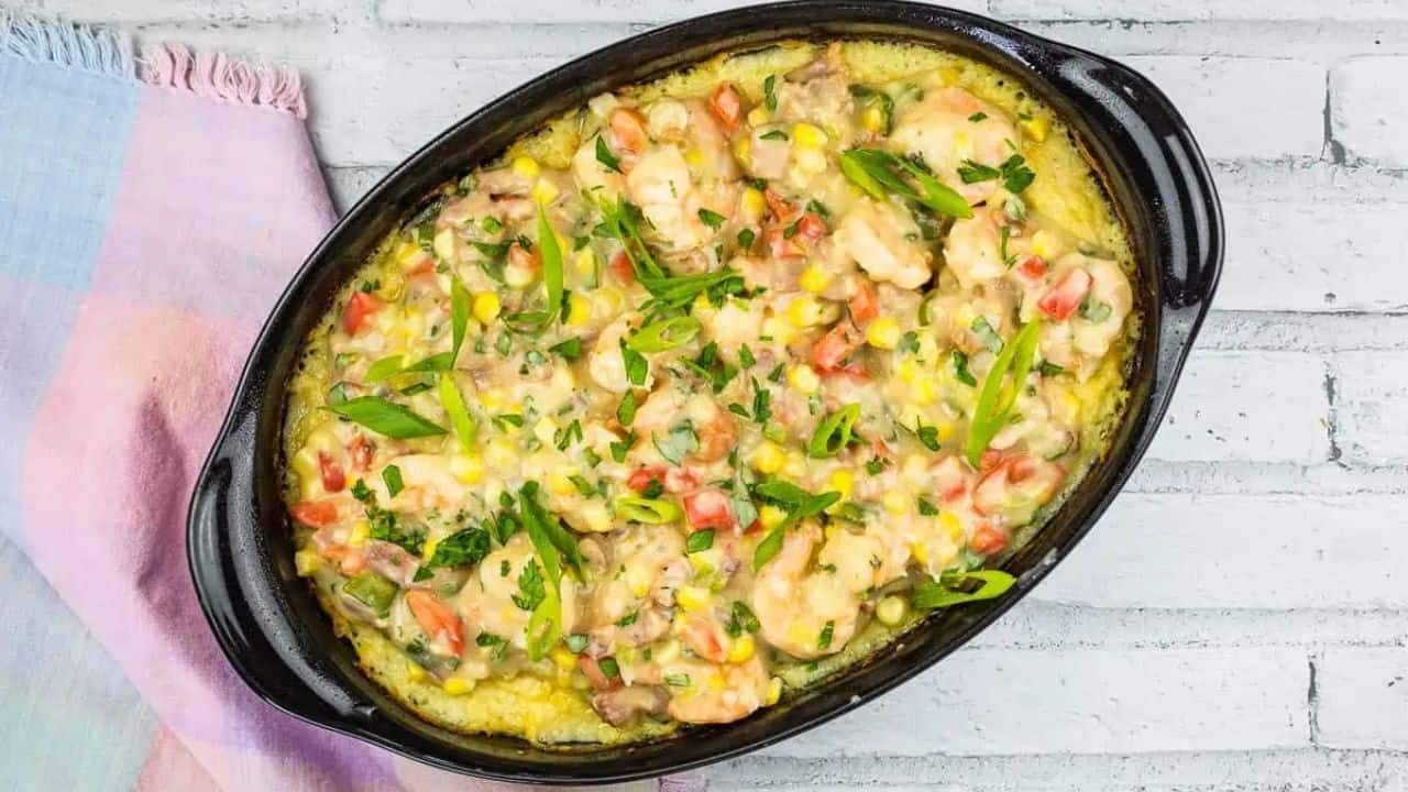 Baked casserole in an oval dish, topped with shrimp, chopped vegetables, and herbs. A colorful cloth napkin is beside the dish, set against a white brick surface.