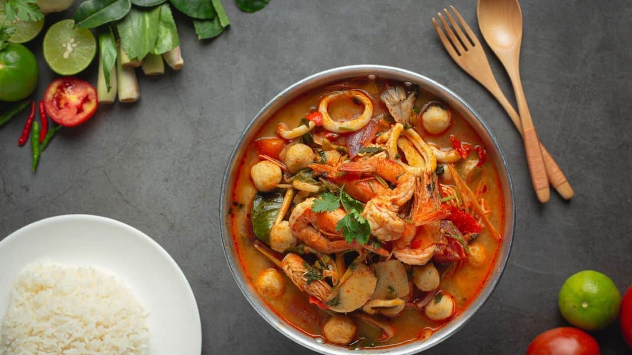 Bowl of seafood soup with vegetables and spices next to a plate of rice, surrounded by fresh ingredients and wooden utensils on a gray surface.