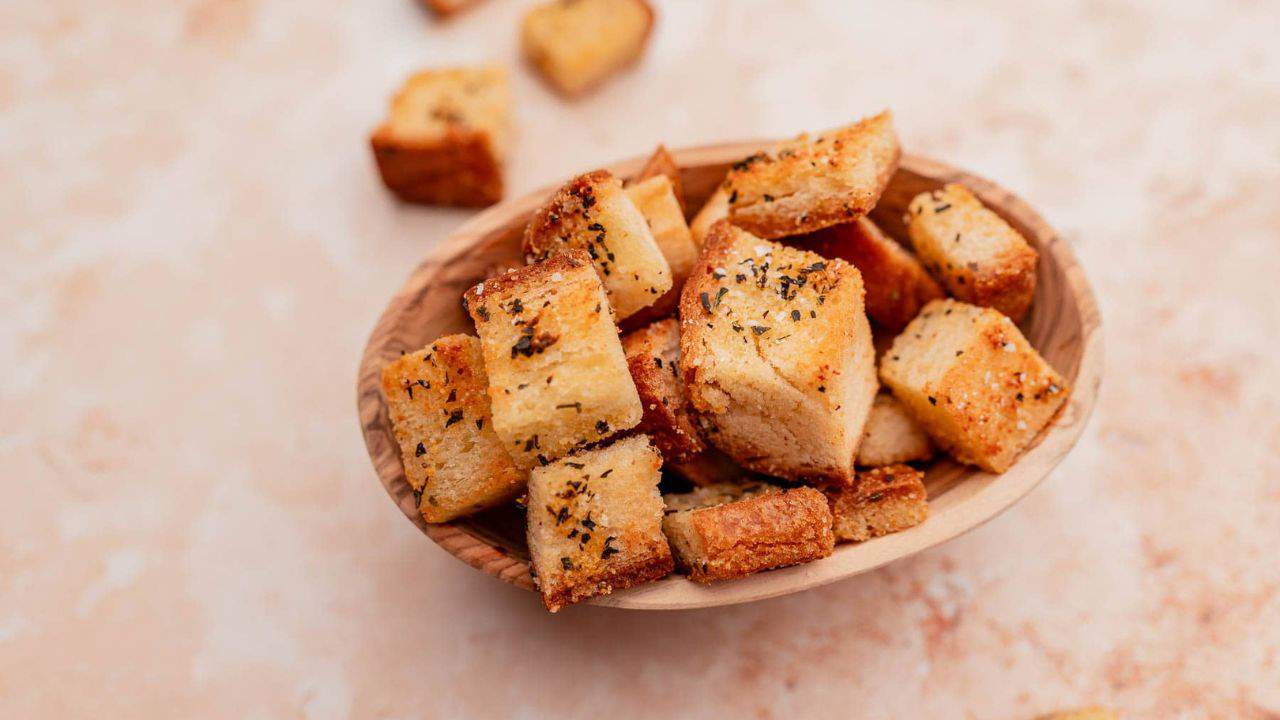A wooden bowl filled with seasoned croutons on a light beige surface, with a few croutons scattered around.