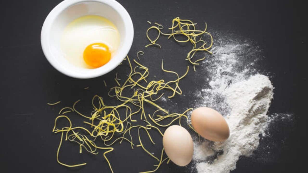 A bowl with a cracked egg, raw pasta, two whole eggs, and flour on a dark surface.