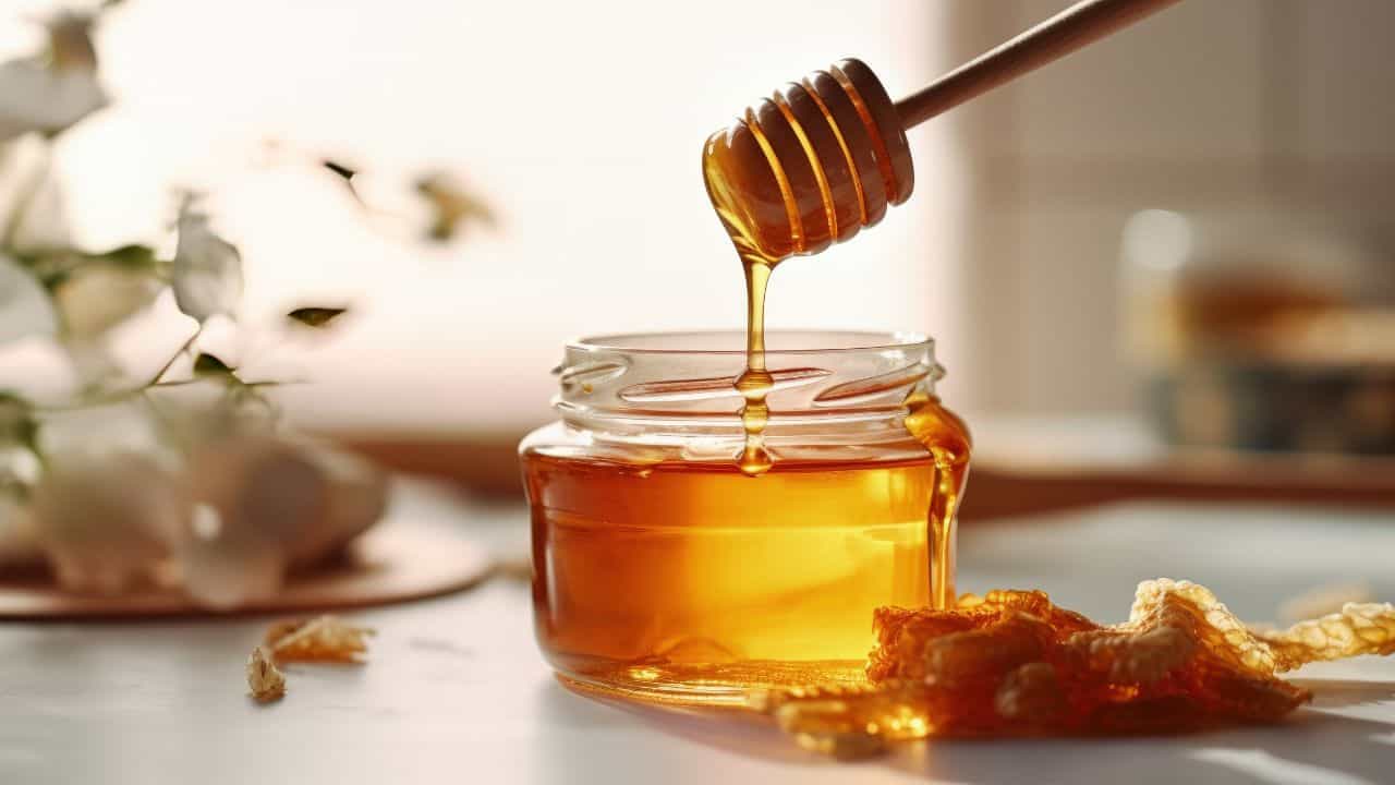A wooden honey dipper drizzles honey into a glass jar on a white surface, with honeycomb pieces nearby. Soft-focus flowers are in the background.