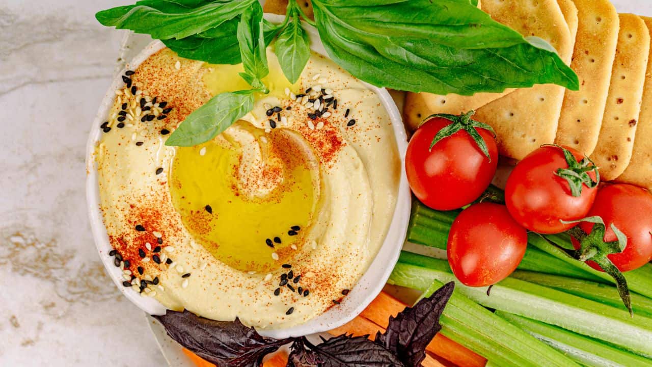 A bowl of hummus garnished with olive oil, sesame seeds, and paprika, surrounded by fresh basil, tomatoes, sliced vegetables, and crackers.