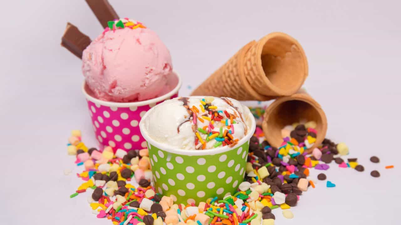 Two polka dot cups of ice cream with sprinkles and chocolate pieces, surrounded by scattered waffle cones and candy toppings on a white background.