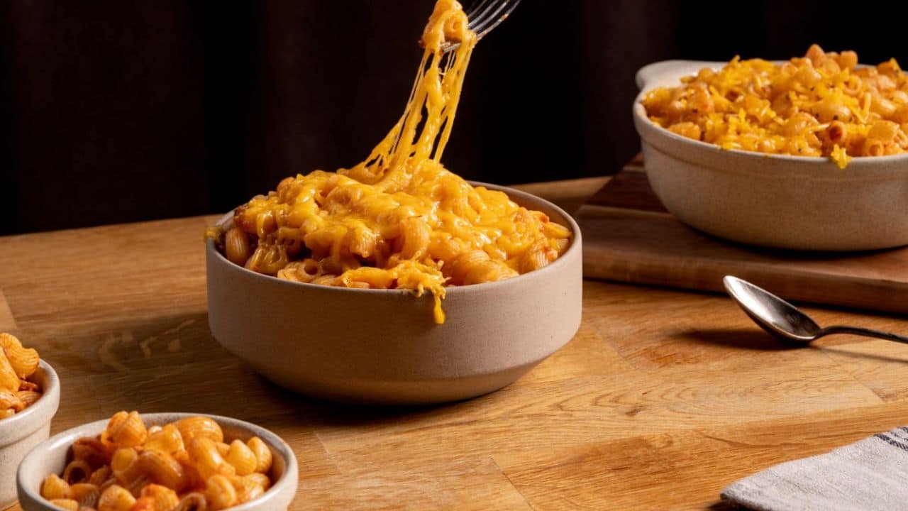 A fork lifts a cheesy portion of macaroni from a bowl on a wooden table. Another bowl of macaroni sits in the background, with a small bowl and spoon nearby.