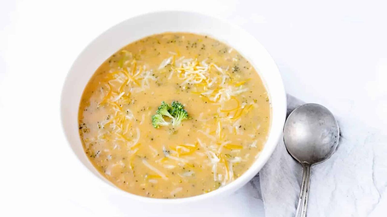 A bowl of creamy broccoli cheese soup garnished with shredded cheese and a small broccoli floret, accompanied by a spoon on a napkin.