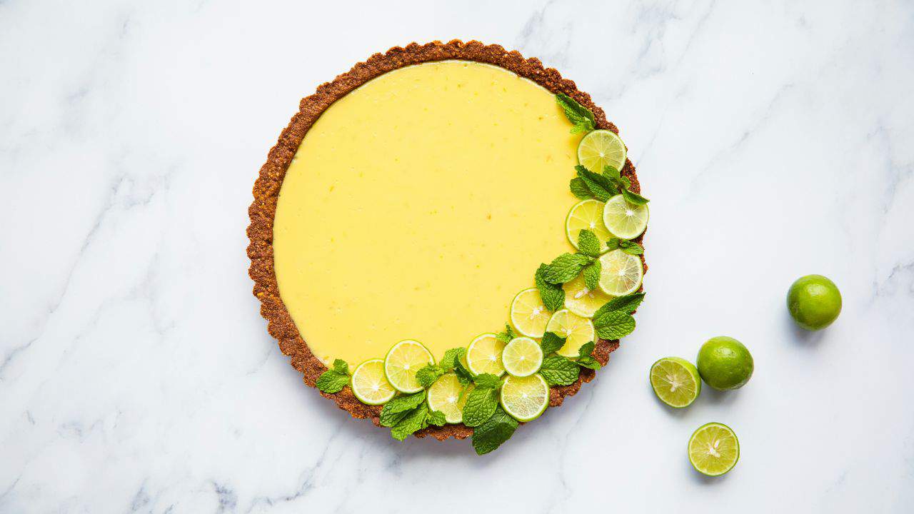 A lime tart with a slice missing, topped with lime slices and mint leaves, placed on a marble surface.