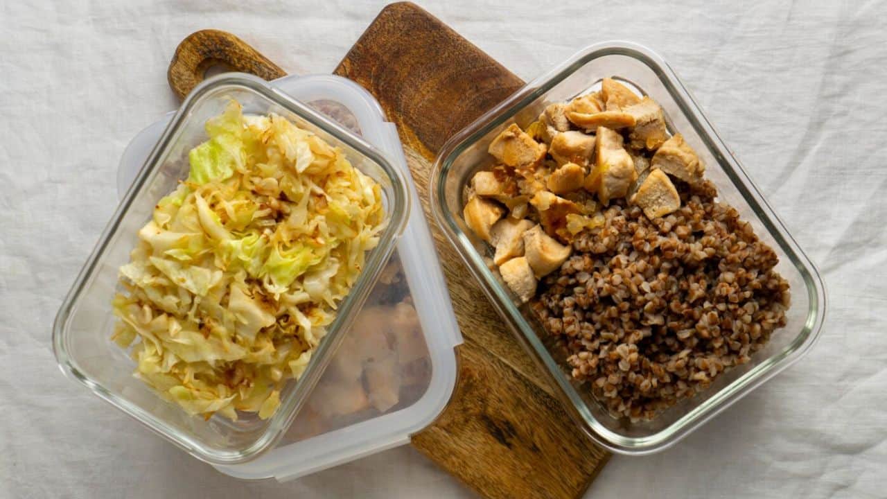 Two glass containers on a wooden board, one with sautéed cabbage, the other with cooked buckwheat and diced chicken.