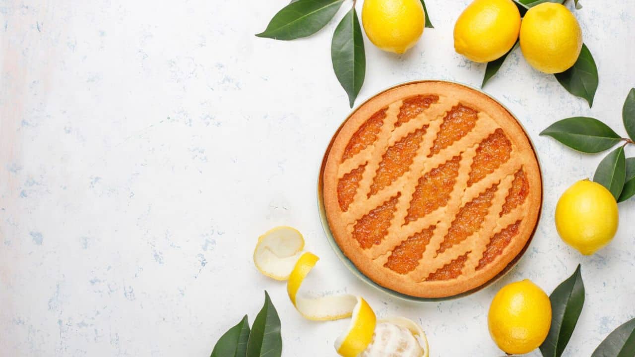 Lemon pie with a lattice top on a light surface, surrounded by whole lemons and green leaves. A lemon peel is also visible.