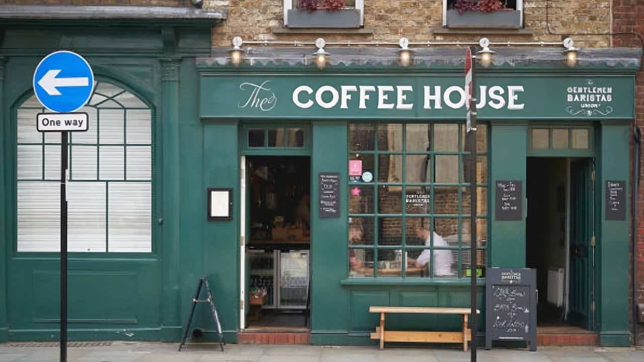 Green coffee shop exterior with signage reading "The Coffee House." A one-way street sign is visible. Two people are seen through the window. Benches and a chalkboard stand outside.
