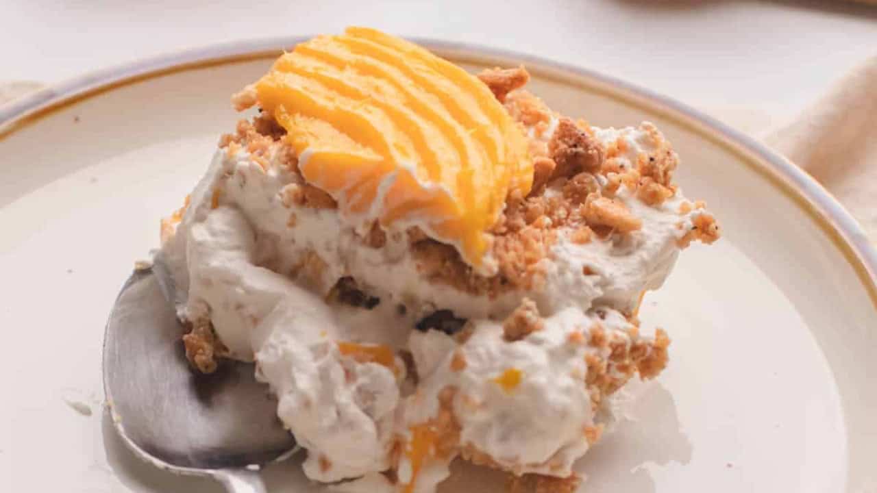 A slice of creamy dessert with crumble topping and sliced ripe mango on a plate, served with a spoon.