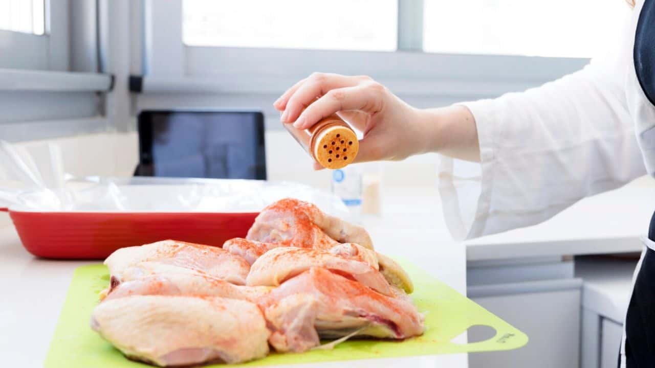 Person seasoning raw chicken pieces on a green cutting board in a kitchen.