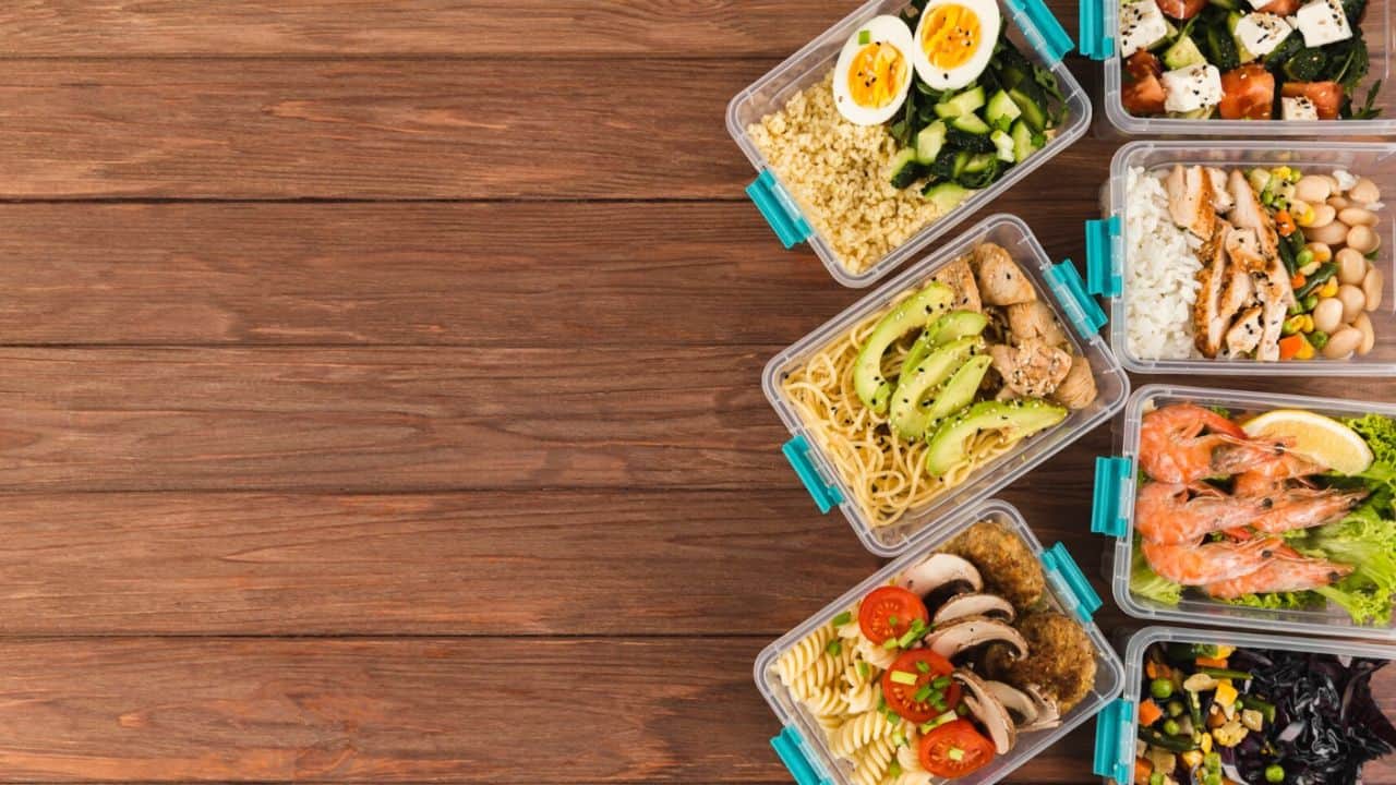 A variety of meal prep containers with different foods, such as shrimp, chicken, pasta, and salad, are arranged on a wooden surface.