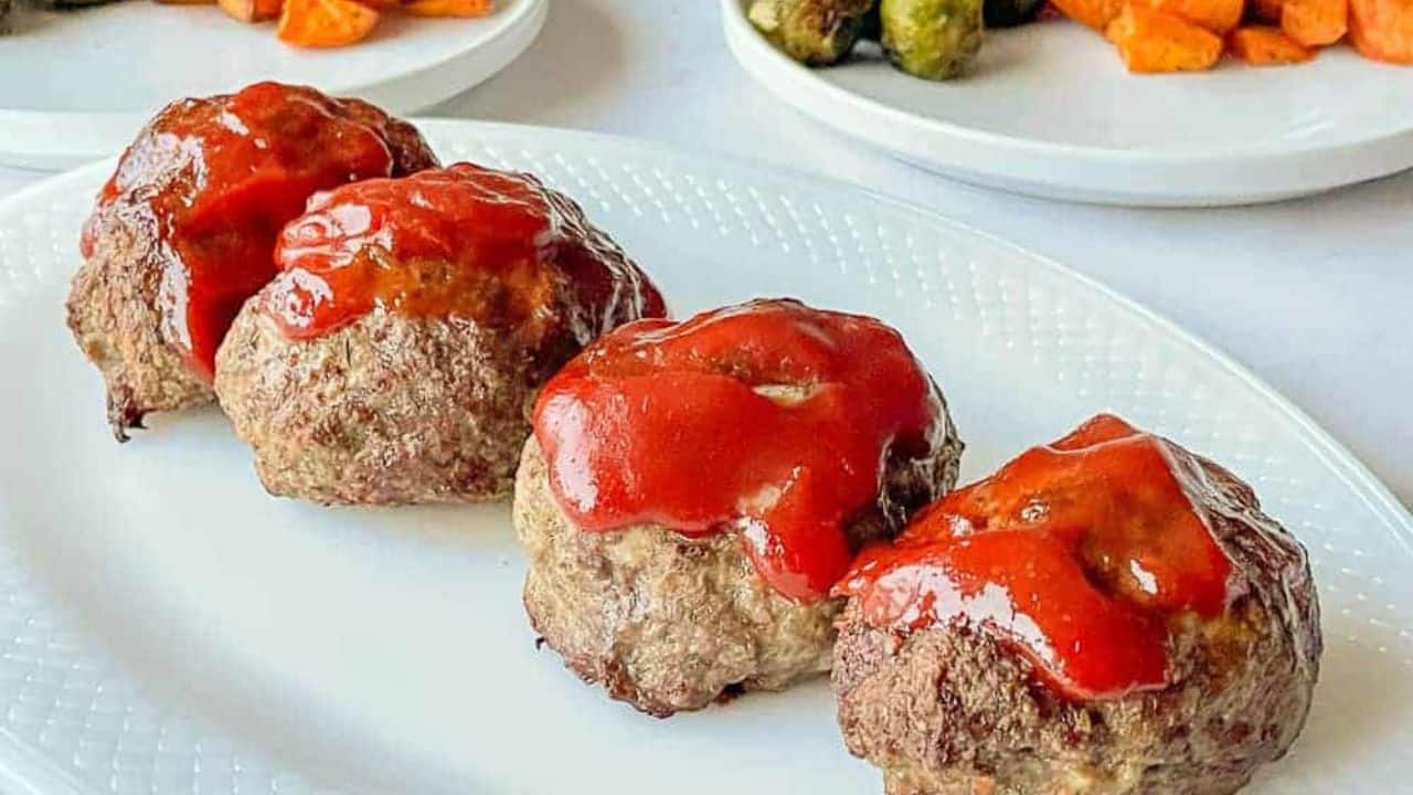 Four meatloaves topped with red sauce are arranged on a white plate. Plates with vegetables are in the background.