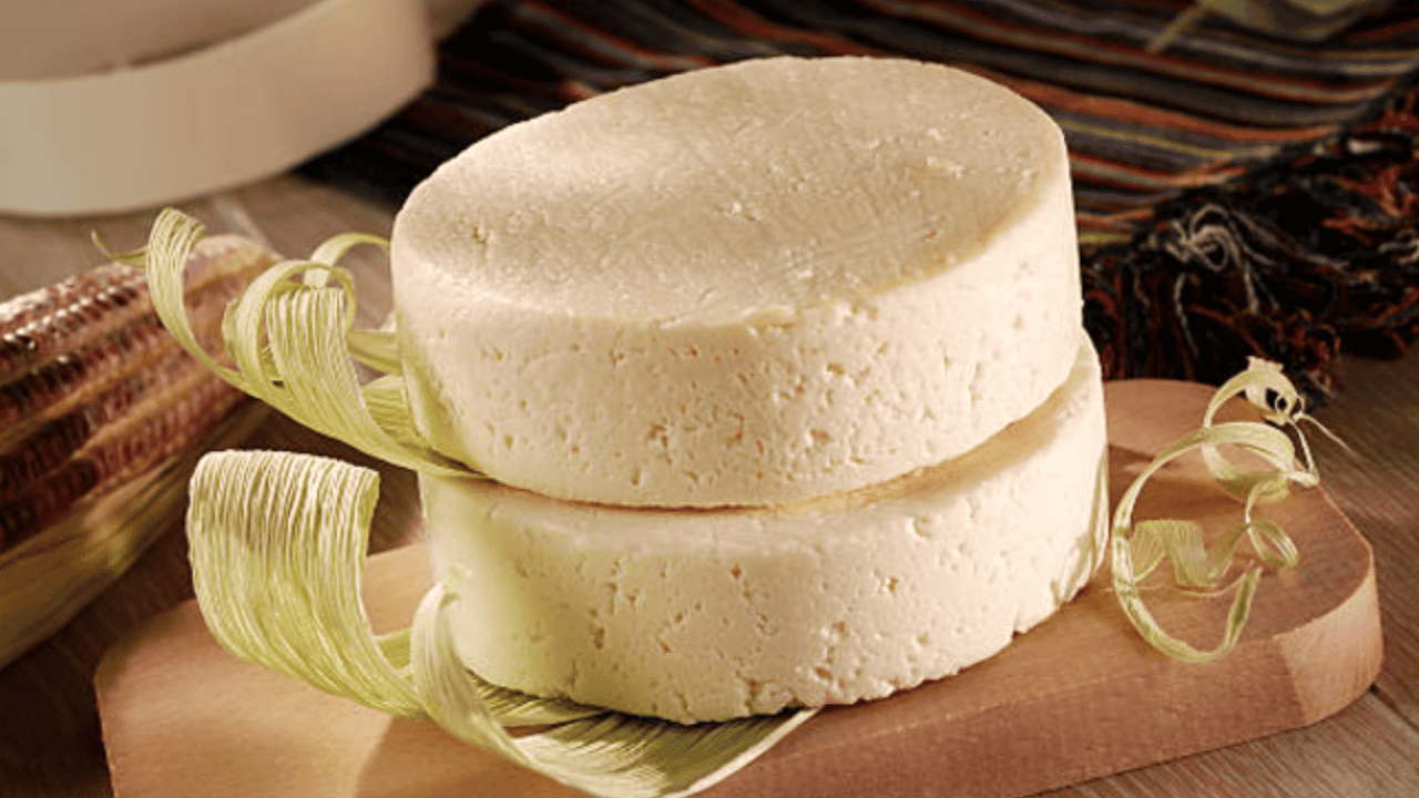 Two round blocks of cheese on a wooden board, surrounded by corn husks, with a striped cloth in the background.