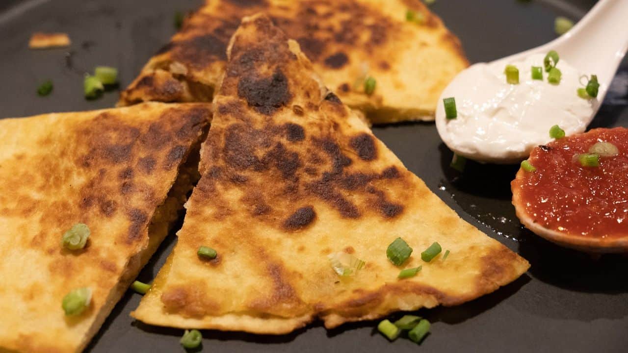 Triangular quesadilla slices on a black plate, garnished with chopped green onions. Served with a dollop of sour cream and a spoonful of red salsa.